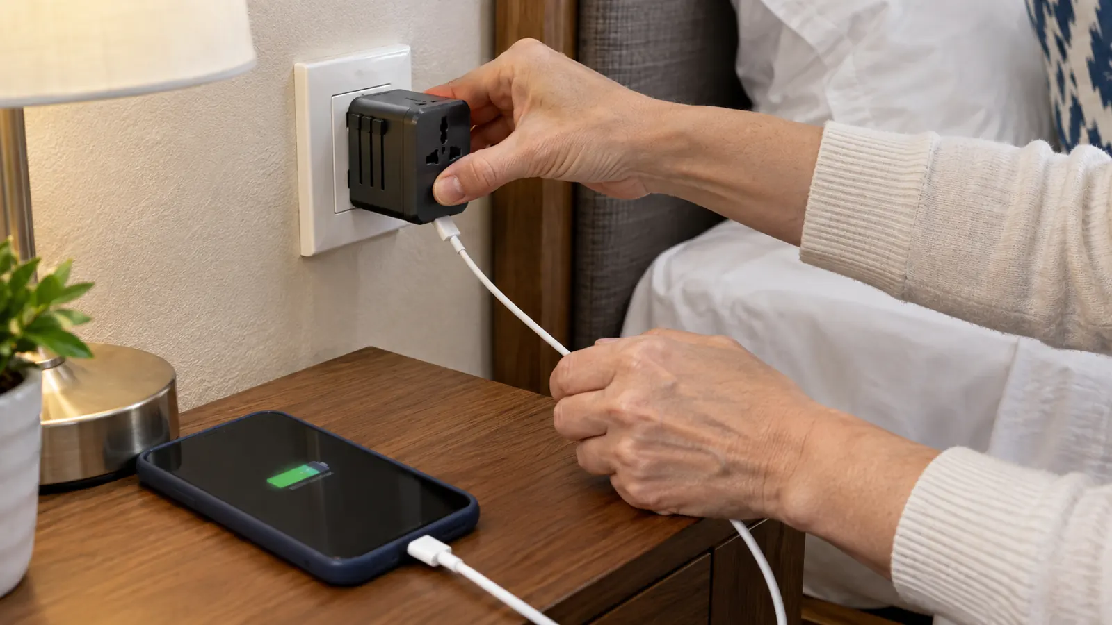 A hand plugging a universal travel adapter into a wall outlet beside a hotel bed, a phone cable connected to it