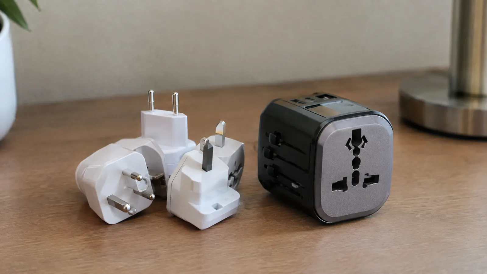 A small pile of single-country plug adapters next to one universal travel adapter on a hotel desk
