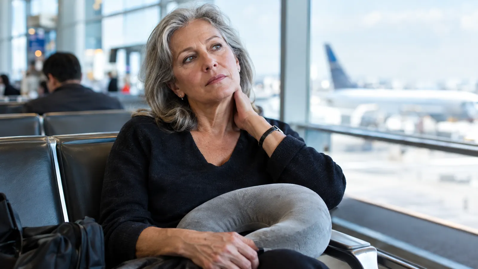 A woman in her sixties sitting in an airport seat, massaging her neck with one hand, a memory foam neck pillow resting on her lap